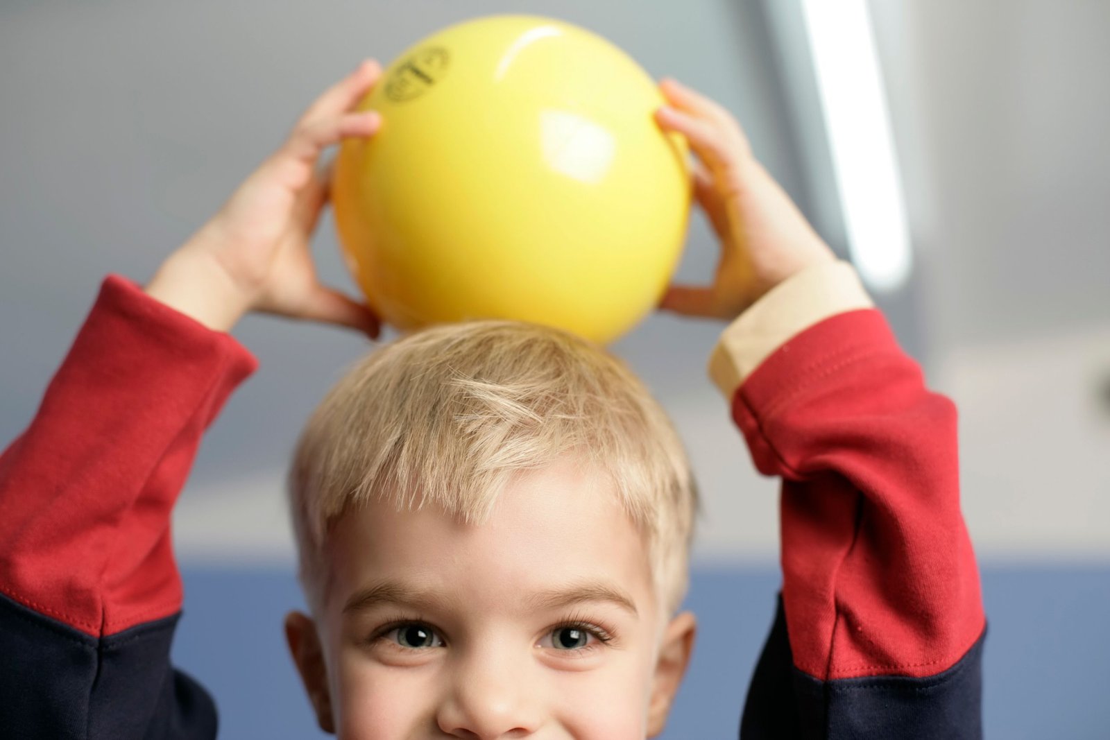 A cheerful child holding a yellow ball above their head, indoors.